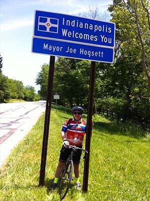 Cyclist Mike Blacconiere stands under Indianapolis welcome sign en route to PurpleStride, the walk to end pancreatic cancer
