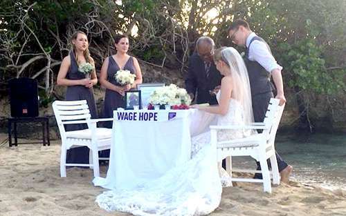 After beachside wedding ceremony, couple sign marriage license at table with Wage Hope banner supporting the cause.