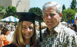 Caucasian female college student at graduation, holding flowers and smiling next to her dad.