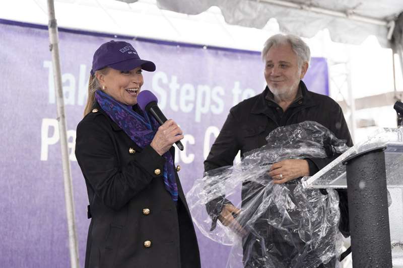 Lisa Swayze holding a microphone and Franke Previte looking at her while both on stage at PanCAN PurpleStride 2023.