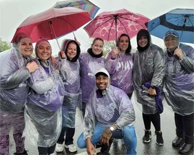 Seven women standing in rain ponchos and one man in a rain poncho posed crouching below them pose for a picture at PanCAN PurpleStride.